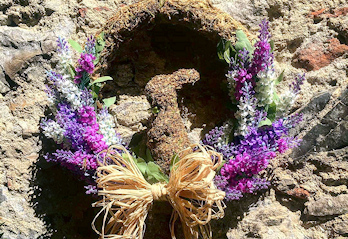 Composizione decorativa naturale con fiori viola, spighe e dettagli intrecciati davanti a una parete in pietra